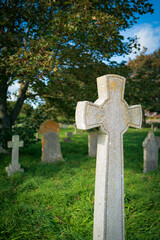 Vertical aspect of an old crucifix shaped headstone seen in a late summer churchyard in England. The lush grass and various headstones can be seen in the background.