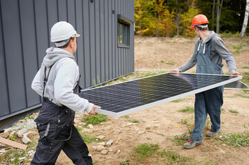 Mounters building solar panel system. Men workers in helmets carrying photovoltaic solar module outdoors. Concept of alternative and renewable energy.