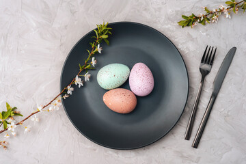 Black plate with Easter eggs, cherry blossom branches and cutlery on textured gray background. Top...