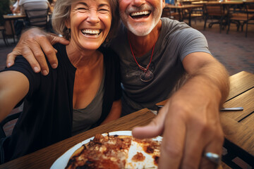 Happy senior old couple have fun eating a pizza together outdoor in traditional italian pizzeria restaurant sitting and talking and laughing. People enjoying food and elderly lifestyle. Tourism