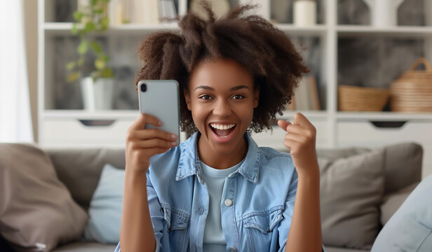 Excited happy young black woman holding smart phone device sitting on sofa at home , Happy satisfied female looking at mobile smartphone screen gesturing yes with clenched fist ,the girl is smiling at
