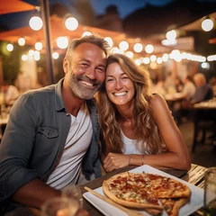 Tuinposter Happy adult cute couple have fun eating a pizza together outdoor in traditional italian pizzeria restaurant sitting and talking and laughing. People enjoying food and travel lifestyle. Tourism © simona
