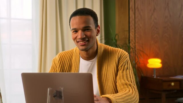 portrait of young Latin man sitting at table working at laptop at home, looks in camera nodding head and smiling in sign of assenting and success. contented and pleased student of online education