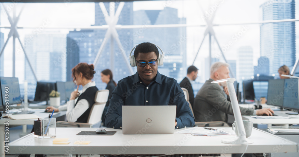 Aufkleber Successful Black Man Wearing Headphones And Starts Typing On Laptop Computer In Skyscraper Office In Downtown. Professional Male Software Developer Working On a New Project In Diverse Tech Startup.