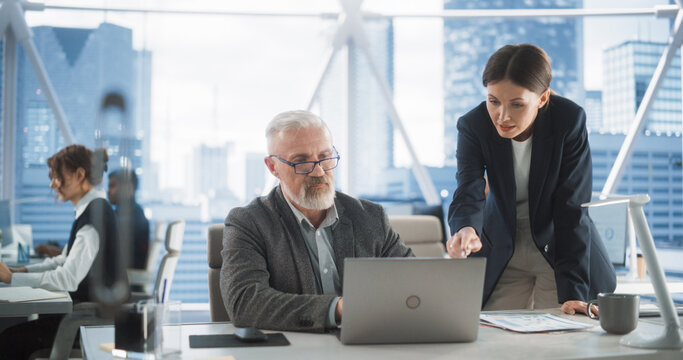 Senior Male Financial Analyst And Female Project Manager Working With Laptop Computer In Modern Downtown Office. Caucasian Man And Woman Looking For Investment Opportunities In Banking or Tech Sectors