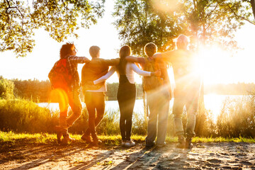 Backlit by the golden sun, a group of five friends shares an embrace, overlooking a peaceful lake. Friends Embracing the Sunset by a Serene Lakeside. High quality photo - obrazy, fototapety, plakaty