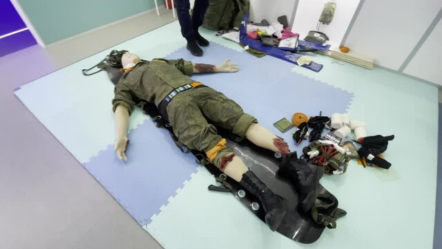 Real time from above view of doll in military uniform with injuries lying on carpet with medical equipment in demonstration room while unrecognizable person in shoes walking on side