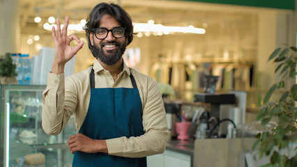 Successful small business owner bearded Indian Arabian man in eyeglasses standing in cafeteria restaurant showing ok gesture smile. Male cafe waiter worker guy with okay sign, good fine coffee service