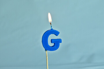 close up on a blue letter G birthday candle with fire on a white background. 