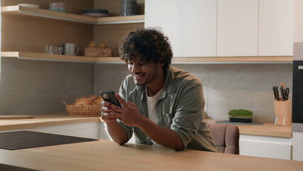 Satisfied Indian male win with phone winning financial prize reward goal achievement with mobile app betting service online victory. Arabian happy man celebrating smartphone success at home kitchen