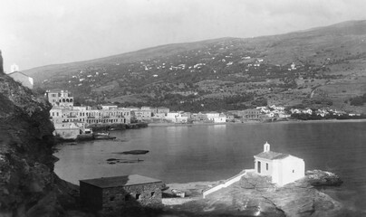 Black and white photo from the island of Andros, Greece, in the 1950s. The edge of the capital town of Andros towards the sea is seen,with small white church in foreground and mountain in the distancw