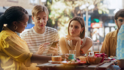 Young Stylish Man Sitting on a Terrace with Multiethnic Friends, Showing Holiday Photos and Social Network Messages on a Smartphone. Young Friends Having Fun Conversations Together in a Cafe