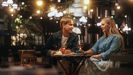 Young Beautiful Couple is Having a Romantic Date at a Cafe at Night. Happy Man and Woman Sitting on a Terrace and Having Fun Conversations During Their Dinner Together in City Center on Weekend - obrazy, fototapety, plakaty