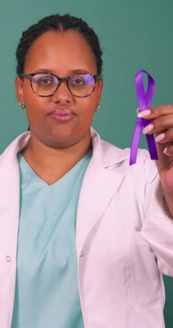 Black female doctor holding purple awareness ribbon, serious in white coat