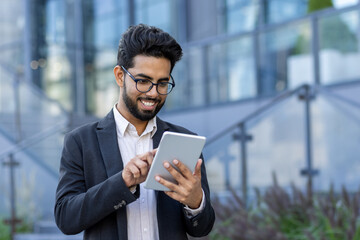 Close-up photo of a smiling young Indian man in a suit sitting near an office building and using a...