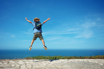 Happy blond kid with backpack jump high excited to see ocean - 755578781