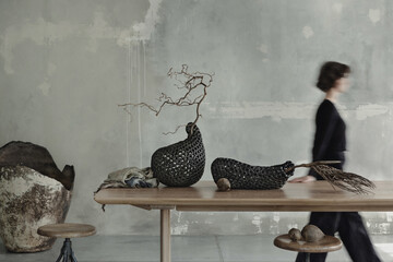 Raw composition of japandi dinning room interior with walking woman, wooden table, gray concrete wall, stylish vase with branch, round stool and personal accessories. Home decor. Template.