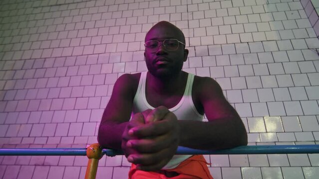 Low angle medium portrait of young Black man wearing white undershirt and eyeglasses leaning on metal railing and looking down at camera, standing against tile wall in room with neon purple light