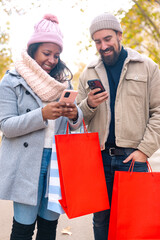 Cute multiethnic couple of man and woman standing holding shopping bags, browsing smartphone apps shopping online