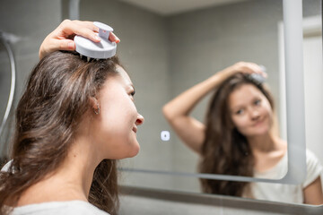 Young woman doing self hair scalp massage with scalp massager or hair brush for hair growth stimulating at home bathroom. Reflected view of the mirror