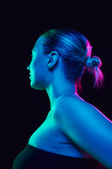 Profile photo of young woman with bare shoulders with bun hairstyle looking away in neon light against black studio background. Concept of spa procedures, cosmetology, skincare products. Ad