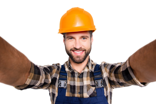 Portrait of handsome cheerful repairer in safety helmet with stubble shooting selfie on smart phone with two hands, wearing shirt and overalls, isolated on grey background
