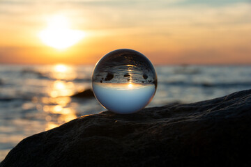 Ball made of glass lies on a stone in which the beach and the sea are reflected - obrazy, fototapety, plakaty