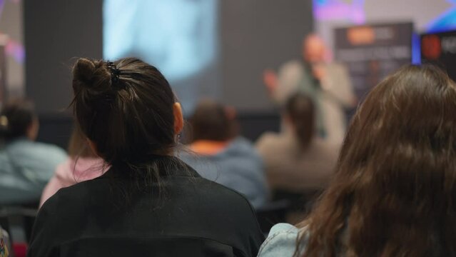 Woman listens attentively to speaker at conference, presenter blurred. Innovative tech presentation captures the attention of young entrepreneurs, foreground focused on audience