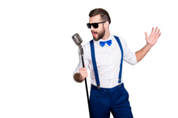 Portrait of handsome attractive singer in blue pants with suspenders and black glasses, singing hit with open mouth in microphone gesture with hand isolated on grey background