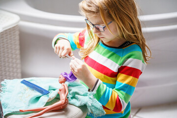 Cute little girl playing with doll at home. Child with eyeglasses making haircut to her toy doll. Indoor creative activities for children