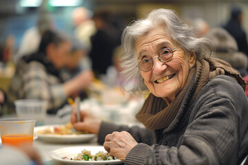 Cheerful elderly homeless woman eats free meal in spacious dining hall. Old lady in need visits soup house to get food. Volunteer care of houseless - obrazy, fototapety, plakaty