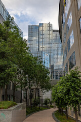 Obraz premium BRUSSELS, BELGIUM, June 23, 2023: A peaceful courtyard with lush greenery nestled between reflective glass office buildings under a partly cloudy sky. Urban Oasis: Green Courtyard Amongst Glass Office