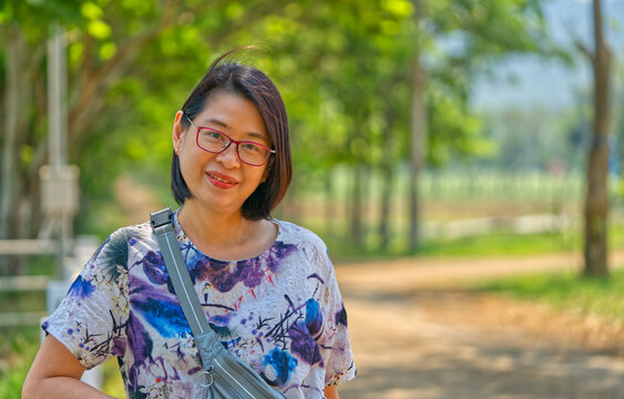 Portrait Asian middle-aged woman on vacation, standing on a farm, smiling and looking at the camera, a woman wearing eyeglasses, and short hair and looks healthy, landscape image with space for copy.