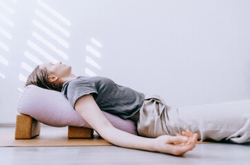 Woman in casual clothing doing yin/restorative yoga with bolster supported by blocks - obrazy, fototapety, plakaty