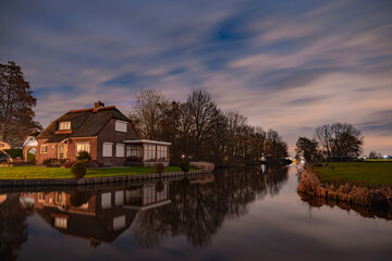 Fototapeta premium Night landscape with starry sky in the Dutch village of Streefkerk. Houses with bright light reflecting in the shine of the water in the autumn season