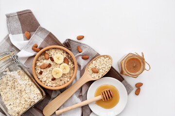 Oatmeal in a wooden bowl with almonds and pieces of banana ready to cook, honey, scattered oat flakes and spoon on white background. View from above. Place for text - 755642901