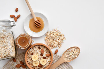 Oatmeal in a wooden bowl with almonds and pieces of banana ready to cook, honey, scattered oat flakes and spoon. View from above. Place for text - 755642914