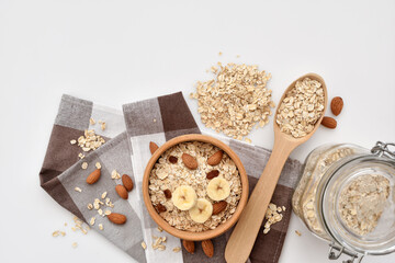 Oatmeal in a wooden bowl with almonds and pieces of banana, scattered oat flakes and with spoon on white background. View from above. Place for text - 755642951