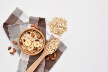Oatmeal in a wooden bowl with almonds and pieces of banana, scattered oat flakes and with spoon on white background. View from above. Place for text - 755642954