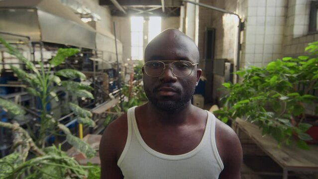 Medium portrait of young African American plant factory worker in undershirt and glasses posing for camera at workplace