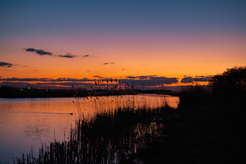 Fototapeta premium Scenic view of beautiful sunset above the lake at spring in the evening with cloudy sky background and reed grass at foreground. Water reflecting in warm color