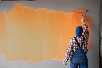 Rear view of painter man painting the wall with paint roller and orange color. Big empty space