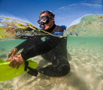 An adventurous snorkeler in a wetsuit is half-submerged in crystal clear waters, with sunlight dancing across the sandy bottom