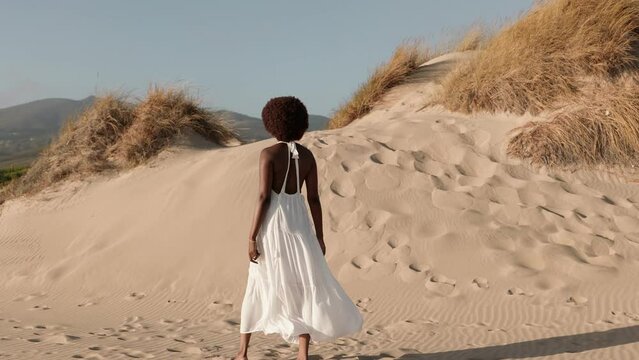 Elegant black woman in a flowing white dress strolls on sandy dunes under a clear sky, embodying tranquility and freedom.