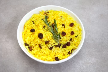 Fotobehang Traditional South African yellow rice, made with turmeric and raisins with rosemary garnish, In round bowl on mottled grey surface  © Aninka