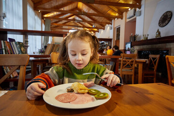 Little girl is having breakfast sitting at wooden table in spacious room