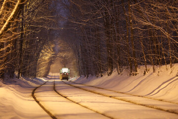 Tram moves on railway on road in park at winter snowy evening, back view