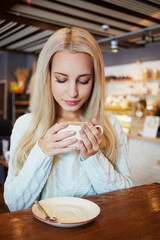 Young blonde woman sits in cafeteria at table, holding cup of coffee in hands