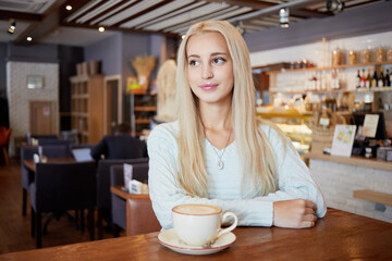 Young blonde woman sits in cafeteria at table with cup of coffee