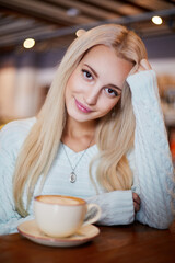 Young smiling blonde woman sits in cafeteria at table with cup of coffee